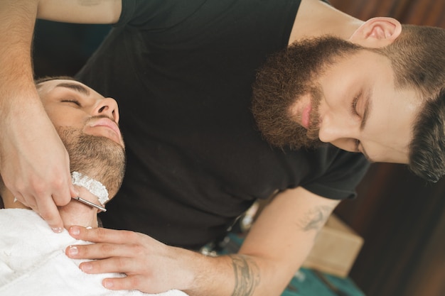 Adora il suo lavoro. barbiere professionista bello che rade il suo cliente in un vecchio modo usando un rasoio