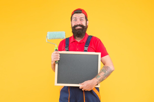 He loves his job. reconstruction advertising. creativity. happy man with paint roller. painter at painting works. hipster with copy space on blackboard. repair and building. craftsman on construction.