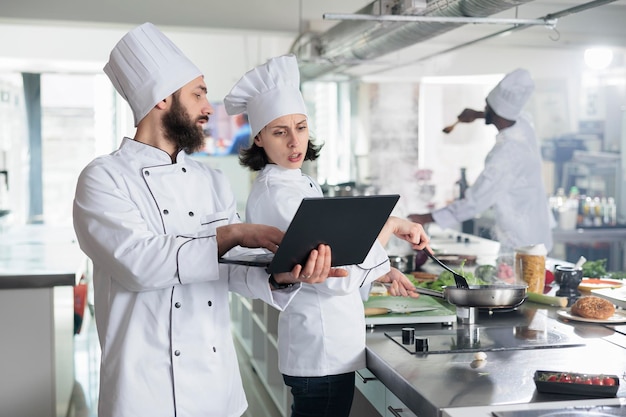 Head chef with modern laptop showing to sous chef recipe for gourmet dish required for gastronomic event held at restaurant. Food industry workers standing in professional kitchen while using computer