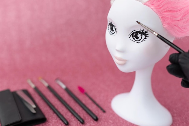 Голова манекена с розовыми волосами для обучения созданию причесокмакияжкоррекция формы бровей на блестящем фоне бокеРука в черной перчаткекистьпинцеттени для глазгубная помада