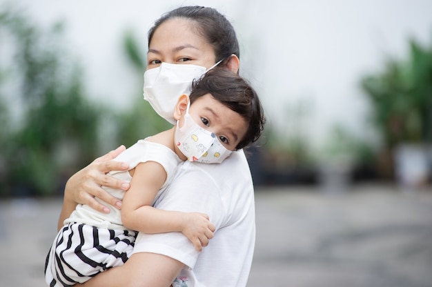 Photo head shot of asian mother wearing white face mask holding her little toddler baby girl who is her daughter wear baby face mask, they look at camera, covid-19 concept