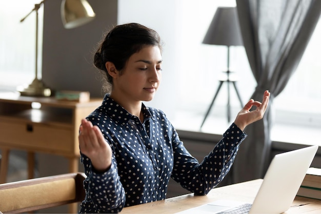 Photo head shot peaceful young indian woman sitting at table making mudra gesture mindful millennial hindu girl freelancer student reducing stress doing breathing exercises alone at home or office