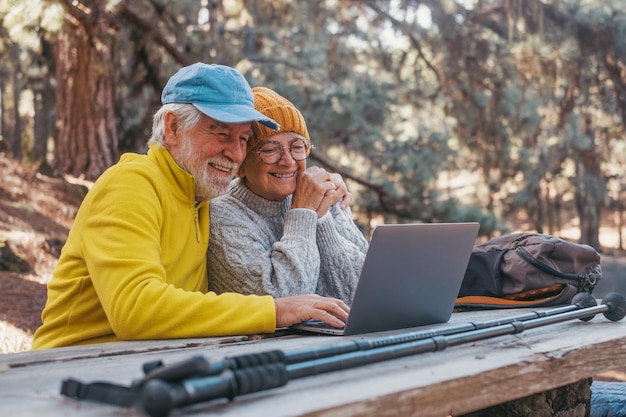 Head shot portrait close up of cute couple of old middle age people using computer pc outdoors sitting at a wooden table in the forest of mountain in nature with trees around them