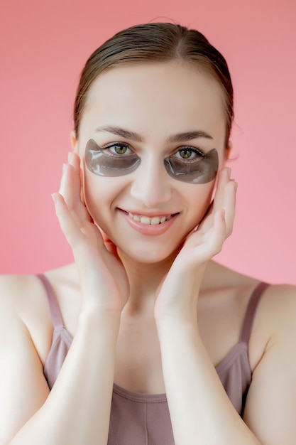 Ritratto di colpo alla testa close up sorridente giovane donna con sotto la maschera di cerotti idratanti occhi guardando la telecamera godendo la procedura di cura della pelle