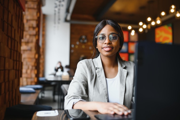 Head shot portrait of happy smiling African American woman sitting at table in cafe, looking at camera, excited female posing, working at computer, doing homework, preparing report in coffee house