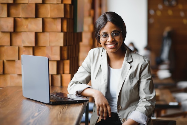 Colpo alla testa ritratto di felice sorridente donna afroamericana seduta al tavolo in caffè guardando la fotocamera eccitata femmina in posa lavorando al computer facendo i compiti preparando il rapporto in caffè