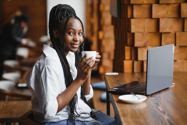 カフェのテーブルに座ってカメラを見て幸せな笑顔のアフリカ系アメリカ人女性のヘッドショットの肖像画は、コーヒーハウスでレポートを準備する宿題をしているコンピューターで働いている女性のポーズに興奮しています