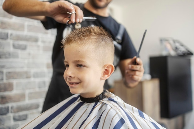 head shot of a smiling little boy sitting in barber shop while hairdresser cutting his hair with scissors