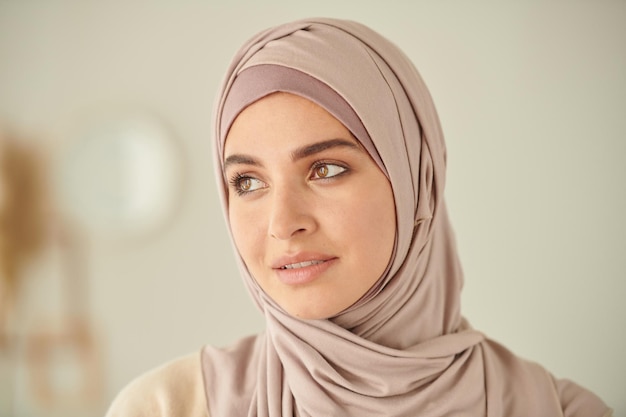 Head and shoulders closeup portrait of young adult muslim woman wearing pale pink hijab looking away