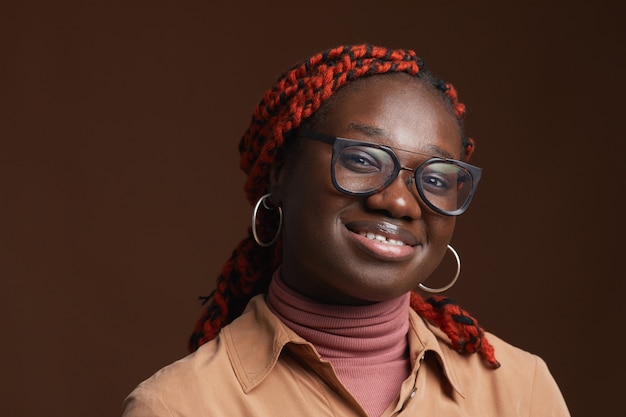 Head and shoulders portrait of contemporary African-American woman wearing glasses and smiling at camera while posing against dark brown background in studio, copy space