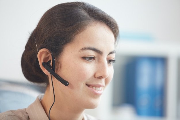 Head and shoulders portrait of smiling young woman wearing headset and talking to customer while working in call center or support service