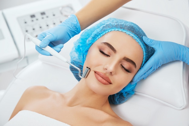 Head and shoulders of woman lying on couch in blue cap in cosmetological clinic with closed eyes. Doctor's hands in blue gloves holding electrode for galvanization near her face