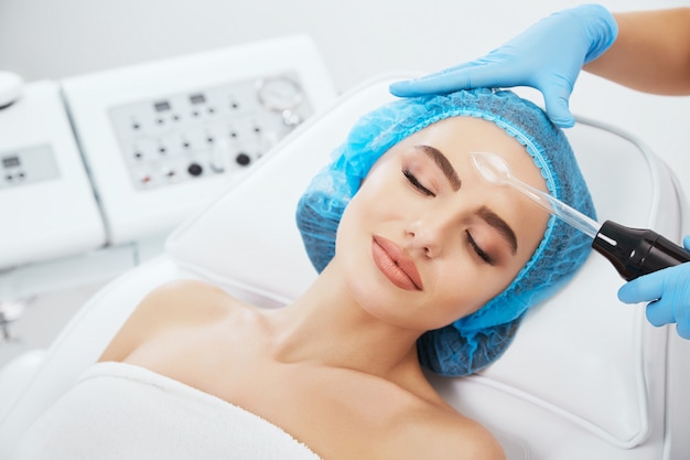 Head and shoulders of woman lying on couch in blue cap in cosmetological clinic with closed eyes and smiling. Doctor holding Darsonvales near her face