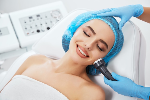 Photo head and shoulders of woman lying on couch in blue cap in cosmetological clinic with closed eyes and smiling. doctor's hands in blue gloves holding ultrasonic phonophoresis near her face