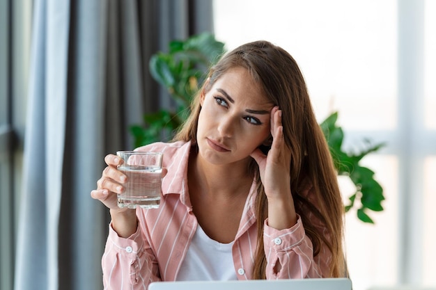 Headache closeup photo of a young woman who is sitting in the\
office getting hydrated drinking water