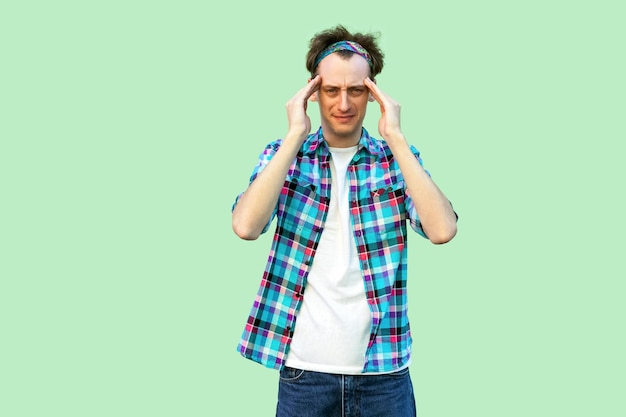 Headache or confuse. Portrait of sad or sick young man in casual blue checkered shirt and headband standing and holding his head and feeling bad. indoor studio shot, isolated on light green background