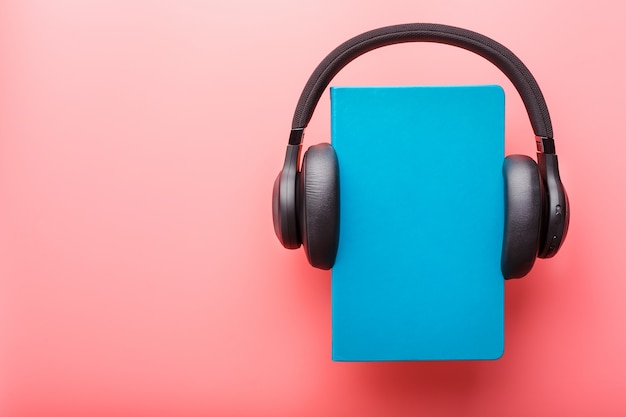 Headphones are worn on a book in a blue hardcover on a pink background, top view.