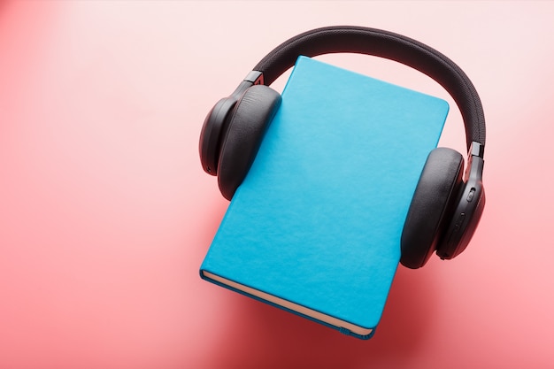 Photo headphones are worn on a book in a blue hardcover on a pink background, top view.