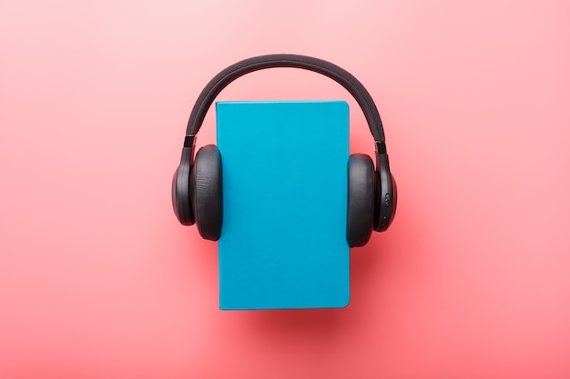 Headphones are worn on a book in a blue hardcover on a pink background, top view.