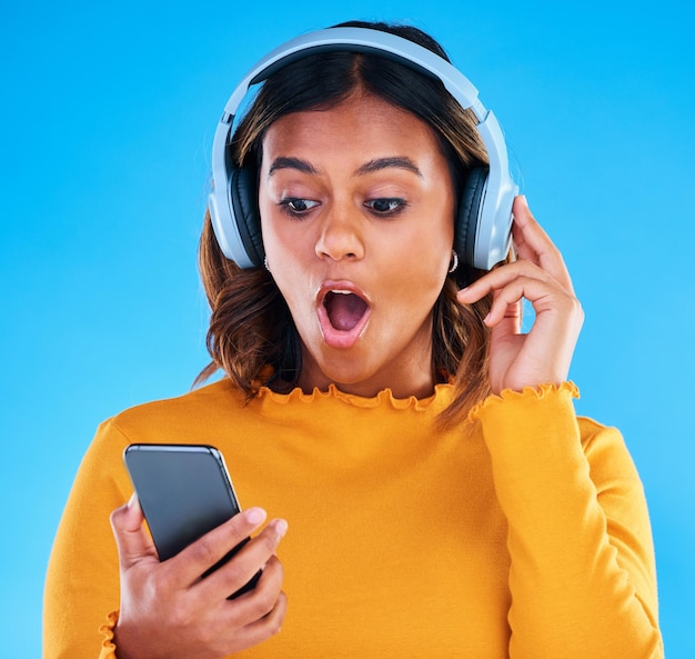 Headphones cellphone and shocked woman in a studio listening to a podcast music or radio surprise technology and female model streaming an album song or playlist with a phone by blue background