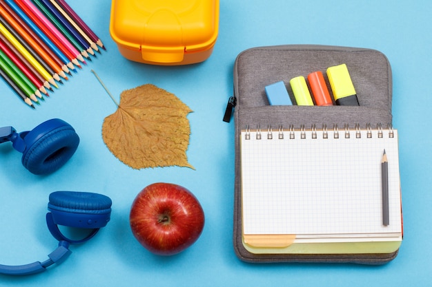 헤드폰, 컬러 연필, 사과, 마른 잎, 도시락, 파란색 배경에 색 펠트 펜과 마커가 있는 가방 연필 케이스에 열린 운동 책. 평면도. 학교 개념으로 돌아가기. 학용품