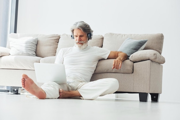 In headphones Sits near sofa Senior stylish modern man with grey hair and beard indoors