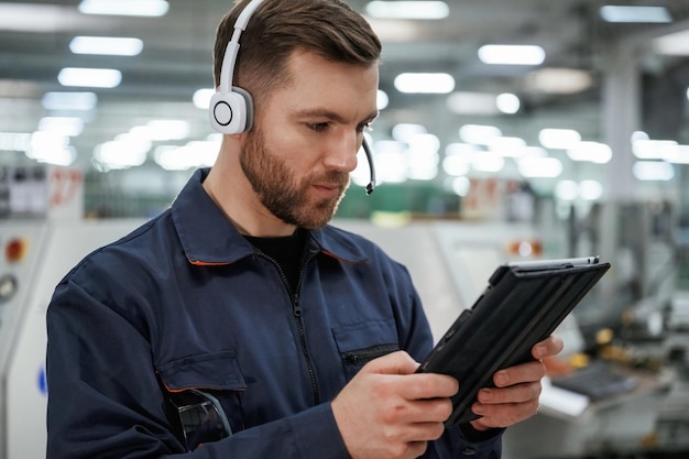 Photo in headphones with tablet factory worker is indoors with hard hat