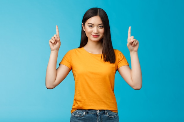 Attenzione, guarda un fantastico promo. donna asiatica sicura di bell'aspetto assertiva che mostra la pubblicità dell'amico, alza il dito indice che punta in alto, macchina fotografica sorridente sicura di sé, sta in piedi con lo sfondo blu determinato.