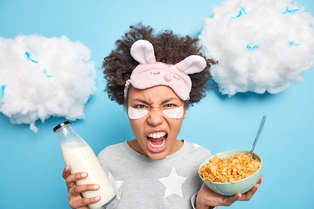 Headshot of angry woman screams loudly keeps mouth opened holds bowl of cereals and bottle of milk has breakfast has bood mood awakes early in morning isolated on blue wall