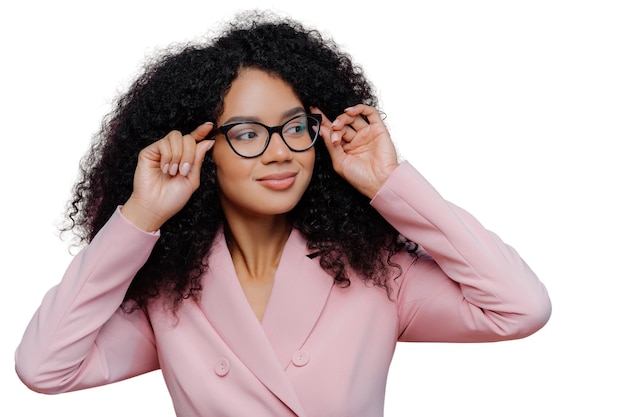 Headshot of attentive female boss keeps hands on frame of glasses looks pensively away wears rosy formal suit has curly Afro hairstyle poses against purple background People business ethnicty