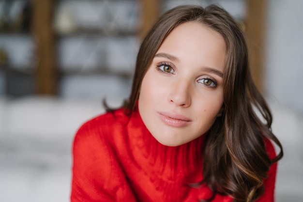 Headshot of attractive healthy European woman with dark hair and soft skin looks directly at camera wears warm red sweater models indoor People and lifestyle concept Domestic atmosphere