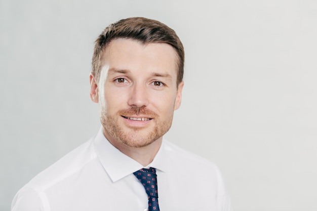 Headshot of attractive male entrepreneur with bristle dressed in formal white shirt and tie looks positively at camera rejoces successful work in office poses alone against white background