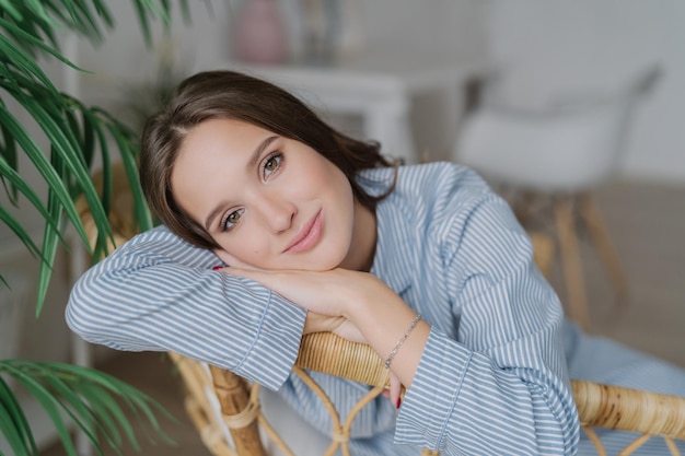 Headshot of brunette young woman with healthy skin and make up leans on hands feels lonely while being at home poses against blurred backround Relaxed girlfriend in striped blue costume