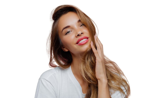 Headshot of carefree relaxed feminine lady tilts head left touches her long wavy hair wears lipstick and makeup prepares for special event in her life being professional photo model stands indoor