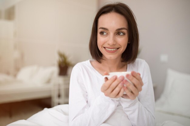 Photo headshot of cheerful young woman drinking herbal tea in bed