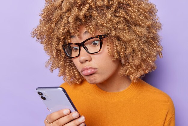 Headshot of curly haired sad worried woman focused at
smartphone feels puzzled reads bad news in internet wears big
transparent eyeglasses and casual orange jumper isolated over
purple background