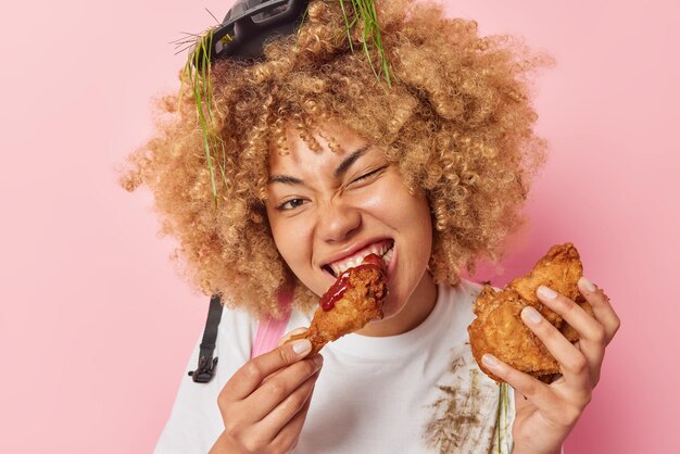 Headshot of curly haired woman eats appetizing fried nuggets\
with ketchup enjoys eating cheat meal wears helmet with stuck grass\
white dirty t shirt isolated over pink background unhealthy\
nutrition