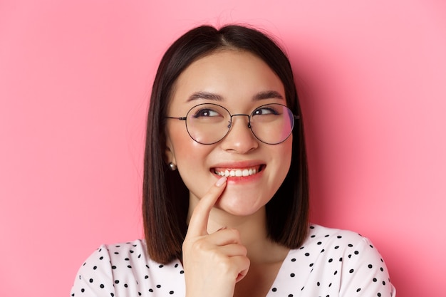 Headshot of cute asian woman in trendy glasses smiling, having an idea, thinking and looking at upper left corner, pink.