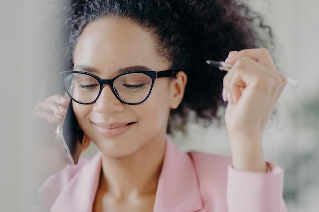 Headshot of delighted female director holds pen in hand wears\
optical eyeglasses has gentle smile makes call via modern cell\
phone dressed in pink elegant costume focused down with\
shyness