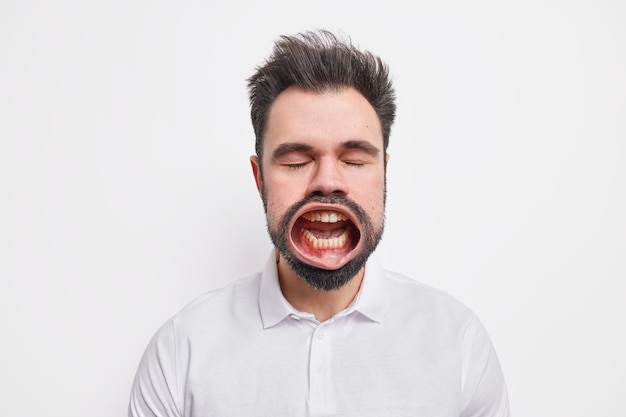 Headshot of furious mad bearded man stands with eyes shut keeps mouth opened screams loudly expresses anger dressed in casual shirt 