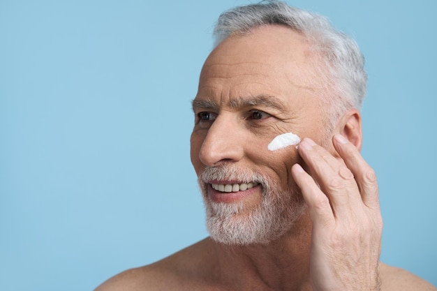 Headshot of handsome senior man applying moisturizing mourishing rejuvenating cream on his face