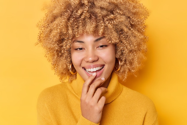 Headshot of happy beautiful woman smiles toohily expresses positive emotions glad to pose in studio for making photo wears casual jumper isolated over yellow background. People and joy concept