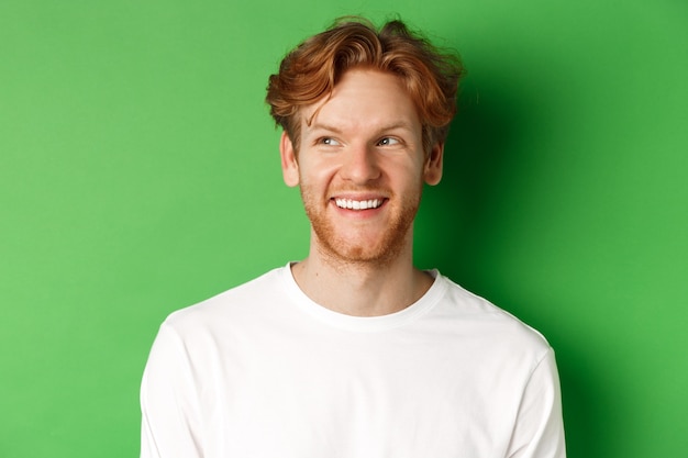 Headshot of happy redhead man with beard wearing white long sleeve, looking left at copy space and smiling, standing over green background.