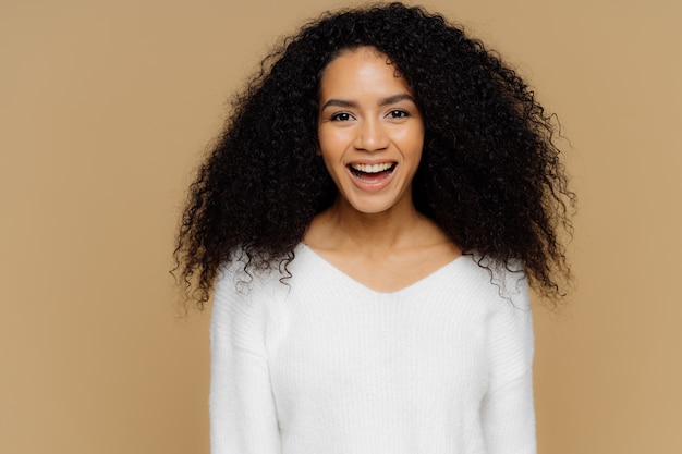 Headshot of healthy positive female model laughs happily, has bushy Afro hair, dressed in white soft jumper