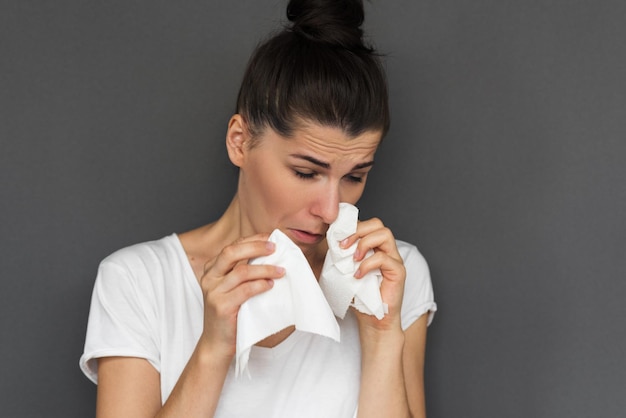 Headshot of ill woman wearing white Tshirt blowing nose into tissue Pretty female have flu virus against gray background Healthy medicine concept Copy space for your text or advertising