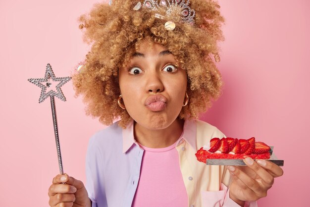 Headshot of lovely surprised young woman pouts lips looks\
stunned at camera eats delicious strawberry cake holds star shaped\
magic wand wears shirt isolated over pink background holiday\
concept