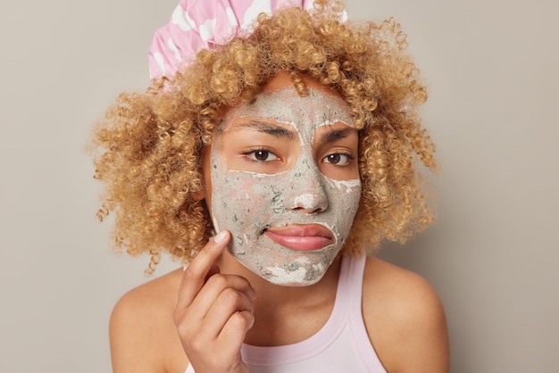 사진 심각한 곱슬머리 여성의 헤드샷은 얼굴에 미용 마스크를 적용하고 피부 상태를 조사하고 목욕 모자를 쓰고 티셔츠는 회색 배경에 고립되어 젊어 보이기 위해 얼굴 치료를 받습니다