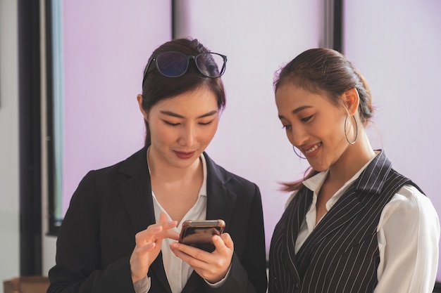 Фото Выстрел в голову двух профессиональных азиатских женщин в деловой повседневной одежде, использующих и смотрящих на смартфон
