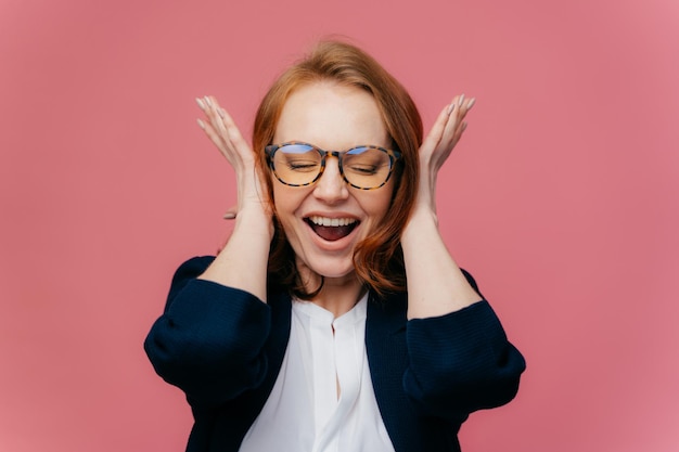 Headshot of overjoyed red haired female entrpereneur comes on corporative party covers ears as hears loud music wears spectacles and elegant suit isolated over pink background expresses happiness