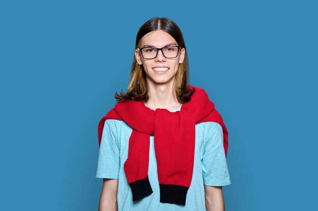 Headshot portrait of cheerful teenage guy looking at camera on blue color background Handsome trendy smiling young male with glasses long hair University college students education youth people